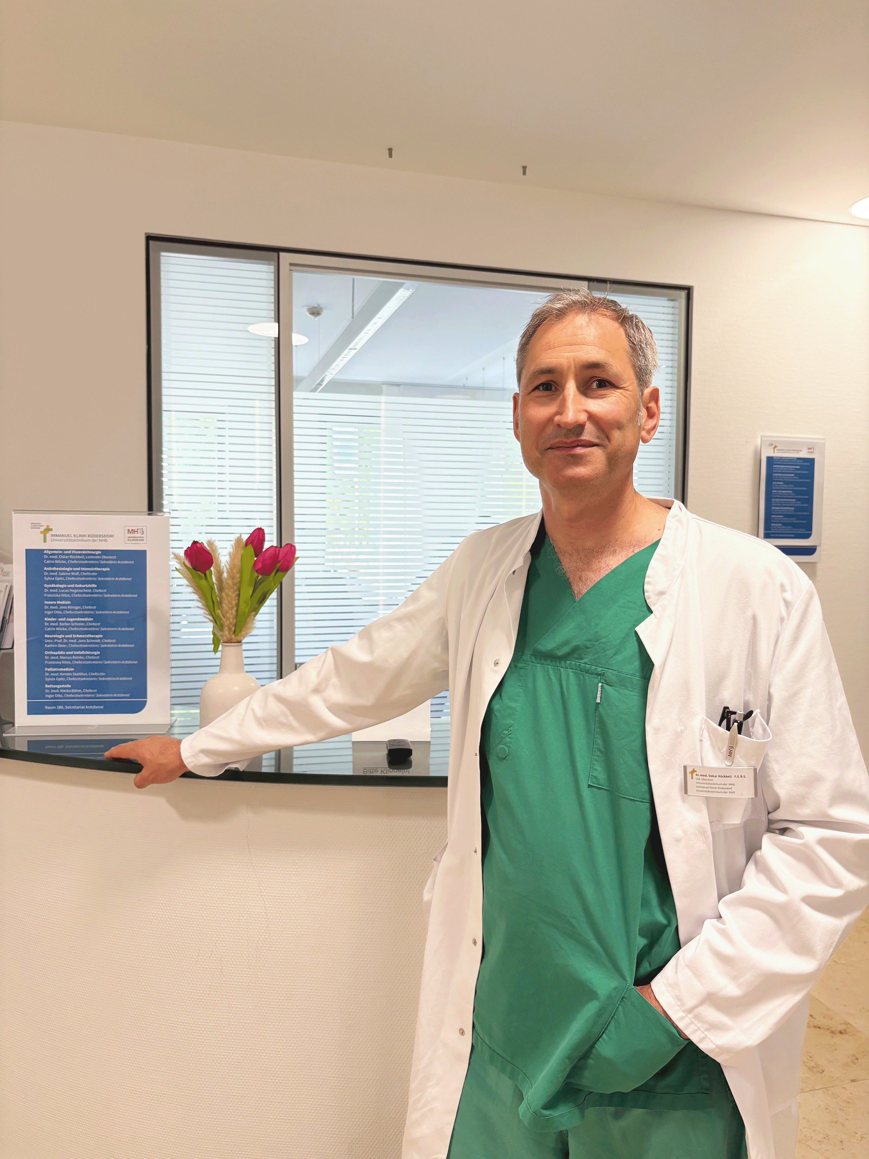 Dr. med. Oskar Rückbeil, kommissarischer Chefarzt der Allgemein- und Viszeralchirurgie, steht in Funktionskleidung am Empfangstresen in der Immanuel Klinik Rüdersdorf beim 18. Rüdersdorfer Chirurgietag zum Thema Inkontinenz und blickt in die Kamera. Dr. med. Oskar Rückbeil, kommissarischer Chefarzt der Allgemein- und Viszeralchirurgie, steht in Funktionskleidung am Empfangstresen in der Immanuel Klinik Rüdersdorf beim 18. Rüdersdorfer Chirurgietag zum Thema Inkontinenz und blickt in die Kamera.
