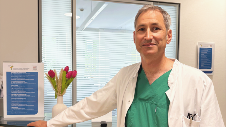 Dr. med. Oskar Rückbeil, kommissarischer Chefarzt der Allgemein- und Viszeralchirurgie, steht in Funktionskleidung am Empfangstresen in der Immanuel Klinik Rüdersdorf beim 18. Rüdersdorfer Chirurgietag zum Thema Inkontinenz und blickt in die Kamera.